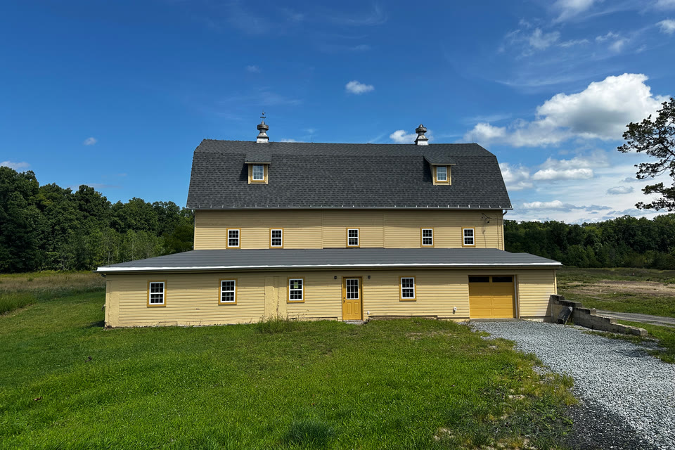 The front of the barn seen from the field, its yellow siding and gray roof against a blue sky.